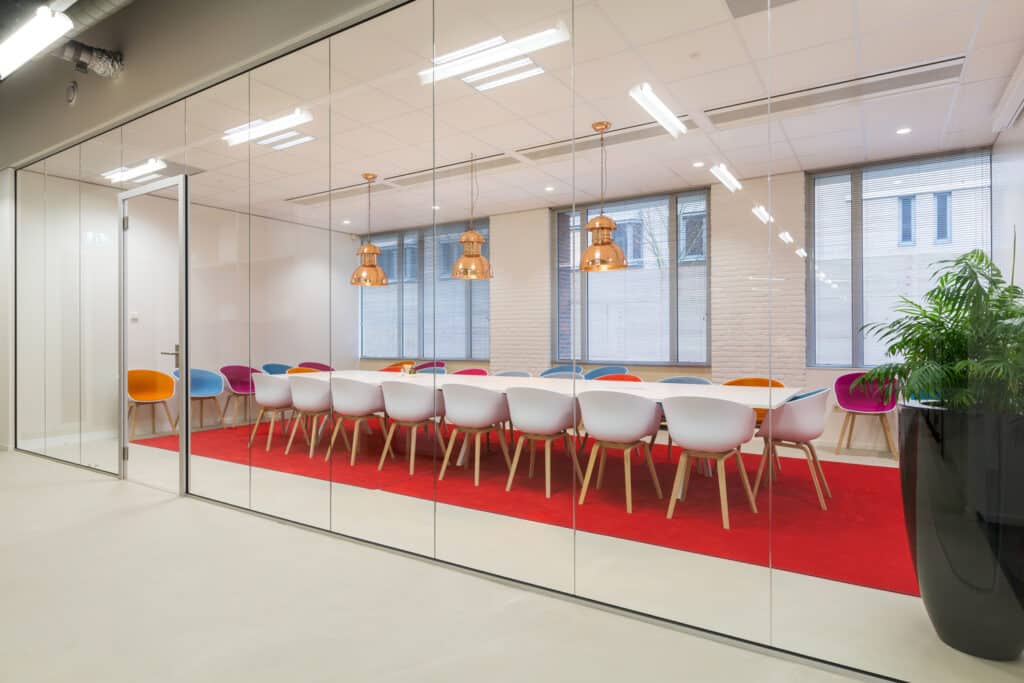 Moderne vergaderruimte met een lange witte tafel, rode loper en transparante slimline glazen wanden.